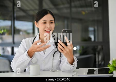 Attraktive junge asiatische Ärztin im weißen Kleid, die ihr Smartphone benutzt, Text liest, E-Mails sendet, mit ihren Freunden während des Bre. Am Schreibtisch chattet Stockfoto