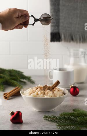 Streuen Sie Zimt über skandinavischen Reisbrei in einer Schüssel, die von Weihnachtskugeln umgeben ist. Frühstück am Silvesterabend. Stockfoto