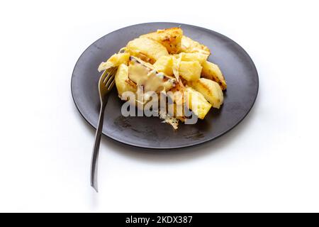 Gebackene Kartoffelkeile, die mit geschmolzenem Käse auf einem schwarzen Teller bedeckt sind, isoliert auf Weiß Stockfoto