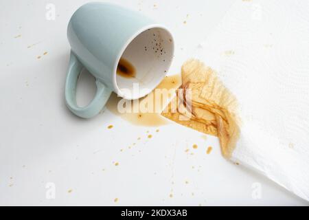 Verschüttete Tasse Kaffee mit einem Stück saugfähigem Papiertuch auf weißem Hintergrund. Nahaufnahme mit weichem Fokus Stockfoto