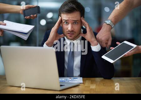 Die Arbeit wird heute etwas zu stressig. Porträt eines gestressten Geschäftsmannes, umgeben von anspruchsvollen Kollegen, während er an einem Schreibtisch sitzt Stockfoto