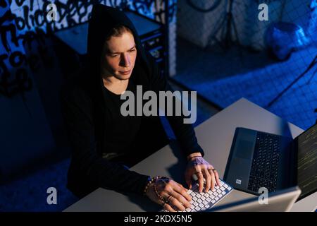 High-Angle-Seitenansicht eines gefährlichen Hackers, der nachts mit einem verstauten Code auf der kabellosen Tastatur arbeitet und Cyber-Sicherheit umgeht. Stockfoto