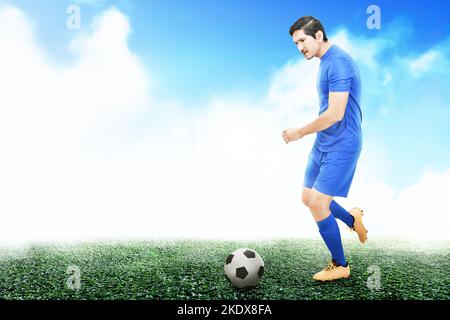 Ein asiatischer Fußballspieler in einem blauen Trikot dribbt den Ball auf dem Fußballfeld Stockfoto