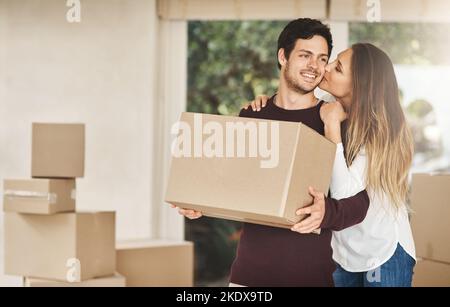 Ein lächelndes junges Paar, das sich küsst, während es am Tag des Umzugens Kisten trägt. Stockfoto