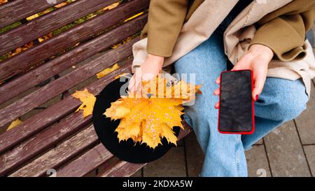 Unkenntliche Frau im Mantel und Schal sitzt auf der Bank. Gesichtsloses Mädchen hält Bouquet von gelben Blättern, Hut und Handy in roter Hülle mit Bildschirm Stockfoto