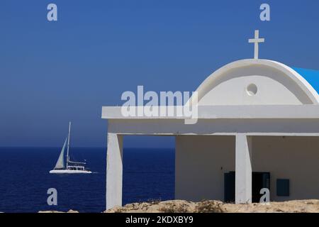 26. Mai 2022, Larnaca, Zypern: Das Segelboot fährt hinter der Agioi Anargyroi-Kirche am Kap Greco vorbei und liegt am Rand einer vertikalen Felsklippe über dem Meer. Es ist den Heiligen Kosmas und Damianos gewidmet. Unterhalb der Kapelle befindet sich eine Höhle mit dem heiligen Wasser der Heiligen. Die Republik Zypern steht an einem historischen und kulturellen Scheideweg zwischen Europa und Asien. Seine Hauptstädte - die Hauptstadt von Nikosia, Limassol, Famagusta und Paphos haben die Einflüsse von Generationen von Eroberern, Pilgern und Reisenden absorbiert und haben eine Atmosphäre, die sowohl kosmopolitisch als auch provinziell ist. (Bild: © Ruaridh Stockfoto