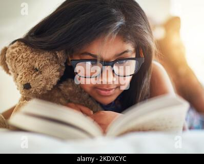 Ich liebe es, mit Teddy an meiner Seite zu lesen. Ein niedliches kleines Mädchen liest ein Buch in ihrem Schlafzimmer. Stockfoto