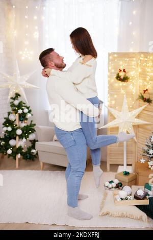 Ein starker Mann hob die Frau in einem weihnachtlich dekorierten Zimmer auf Stockfoto