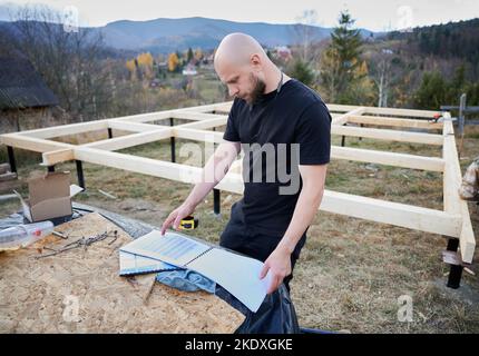 Männlicher Ingenieur, der ein Holzrahmenhaus auf einem Pfahlfundament baut. Baumeister steht auf der Baustelle, mit Baudokumentation, die die Qualität der Arbeit überprüft. Stockfoto