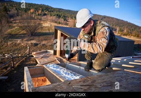 Männlicher Baumeister macht Wärmedämmung auf dem Dach des Holzrahmenhauses. Mann Arbeiter sprühen Polyurethan-Schaum auf dem Dach der zukünftigen Hütte. Bau- und Isolierkonzept. Stockfoto