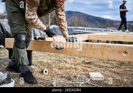 Mann Arbeiter Gebäude Holzrahmenhaus auf Pfahlfundament. Der Zimmermann hämmert mit Hammer einen Bolzen in das Holzwerk. Schreinerei-Konzept. Stockfoto