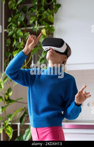 Junge Frau mit Pferdeschwanz in blauem Pullover spielt gerne interessante Spiele in VR-Brillen zu Hause Stockfoto