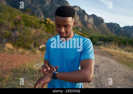 Schwarzer junger afroamerikanischer Erwachsener, der den Timer auf seiner smarten Uhr einstellt, um sich auf seinen Lauf im Freien in den Bergen vorzubereiten. Seine Fitness mit seinem verfolgen Stockfoto