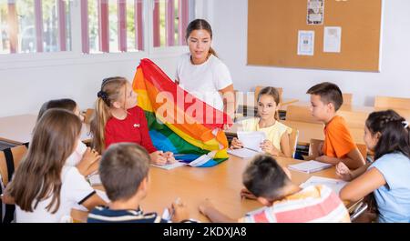Kinder, die im Klassenzimmer sitzen und der Lehrerin zuhören. Sie hält Regenbogenfahne in den Händen und spricht über Minderheiten Stockfoto