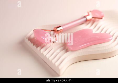 Massageschaber aus Rosenquarz und Gesichtsmassagerolle auf einem geometrischen Podium aus Gips. Anti-Aging-Behandlungen. Gesichtspflege Stockfoto