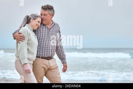 Seniorenpaar, Umarmung und Spaziergang am Strand für Urlaub, Reise oder Liebe in Beziehung miteinander auf mockup. Glücklicher älterer Mann und Frau entspannend Stockfoto