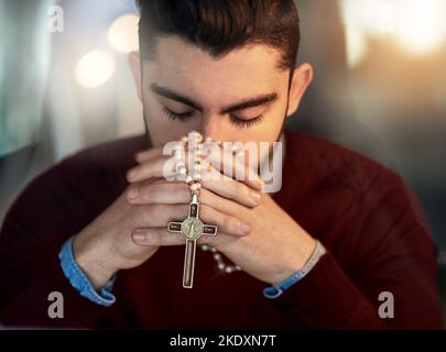 Rufen zu einer höheren Macht. Ein junger Mann, der allein betet, während er einen Rosenkranz hält. Stockfoto