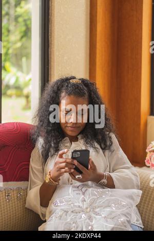 Indische Frau stöbert auf dem Handy, während sie im hellen Wohnzimmer zu Hause auf der Couch sitzt Stockfoto
