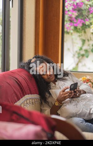 Indische Frau stöbert auf dem Handy, während sie im hellen Wohnzimmer zu Hause auf der Couch sitzt Stockfoto