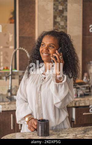 Fokussierte glückliche indische Frau mit schwarzen lockigen Haaren in Weiß tragen Telefongespräche in der hellen Küche zu Hause Stockfoto
