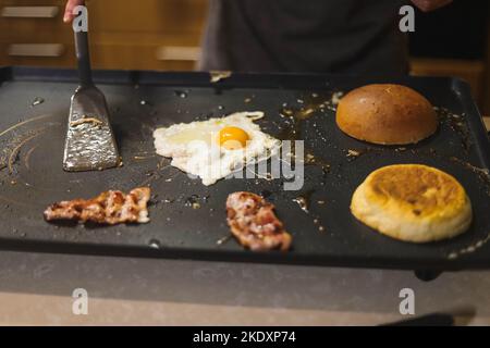 Von oben der Ernte anonymen männlichen Chef in Schürze mit Spatel Bratzutaten für leckere Hamburger auf elektrischem Grill in der Küche Stockfoto