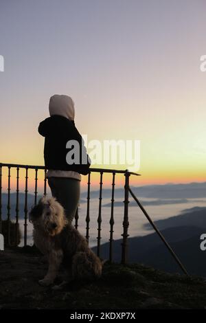 Rückansicht einer nicht erkennbaren Person mit Kapuze auf bewundernde Aussichten mit freundlichem Hund gegen Berge und bewölkten Himmel während des Sonnenuntergangs Stockfoto