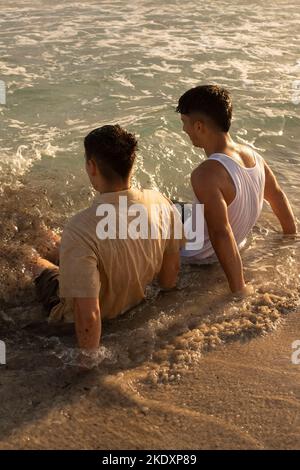 Von oben jungen Jungs in Sommerkleidung sitzen in flachem, winkenden Meerwasser am Strand Stockfoto