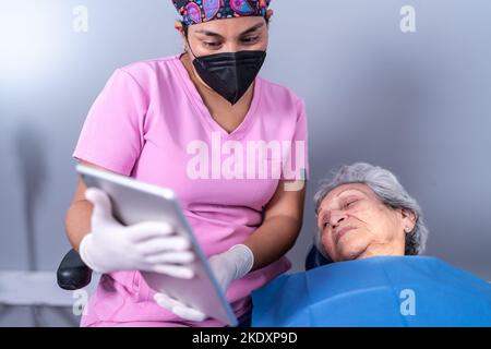 Crop junge ethnische weibliche Zahnarzt in Uniform und medizinische Maske zeigt Bilder auf Tablette für ältere Patienten während der Konsultation in zeitgenössischen Zahn c Stockfoto