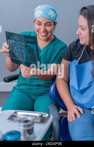 Konzentrierte junge hispanische weibliche Zahnärztin in Uniform und Maske, die dem Patienten ein Röntgenbild zeigt und die Behandlung in einer modernen Zahnklinik erklärt Stockfoto