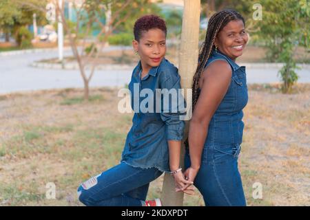 Seitenansicht einer schwarzen Mutter und Tochter in Denim-Kleidung, lächelnd und schauend auf die Kamera, während sie sich Rücken an Rücken stehend und die Hände in der Nähe des Tropica-Rumpfes hält Stockfoto