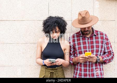 Lateinamerikanisches Paar in legerer Kleidung, das am Wochenende gemeinsam auf Smartphones unterwegs ist, während es an der Stadtmauer steht Stockfoto