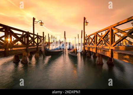 Traditionelle Gondeln, die in der Nähe von hölzernen Kais im San Marco Becken vor dem bewölkten Sonnenuntergang in Venedig, Italien, vertäut sind Stockfoto