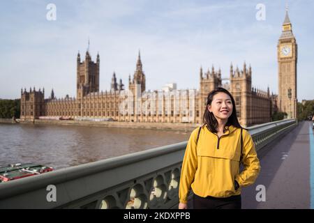 Fröhliche junge asiatische Tourist, die lächelt und weiß wegschaut, steht am Wochenendtag in London auf der Westminster Bridge über der Themse in der Nähe von Big Ben Stockfoto