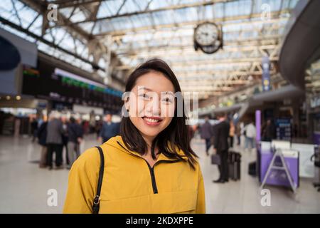 Glückliche junge asiatische Frau mit schwarzem Haar lächelnd und Blick auf die Kamera auf verschwommenem Hintergrund des Bahnhofs in London, Großbritannien Stockfoto