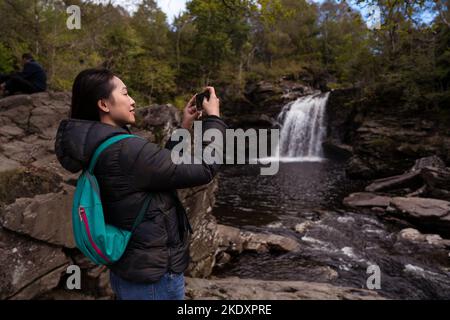 Seitenansicht einer asiatischen Frau in Oberbekleidung mit Rucksack, die am Wochenende in Schottland mit dem Smartphone die Fälle von Falloch fotografiere Stockfoto