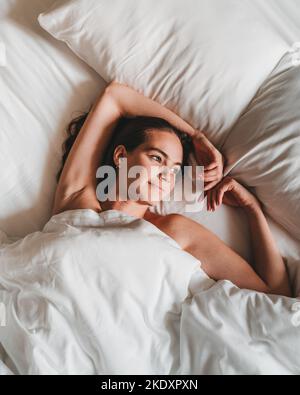 Von oben junge, entspannte Frau, die unter einer weichen weißen Bettdecke im Bett liegt und morgens beim Träumen wegschaut Stockfoto