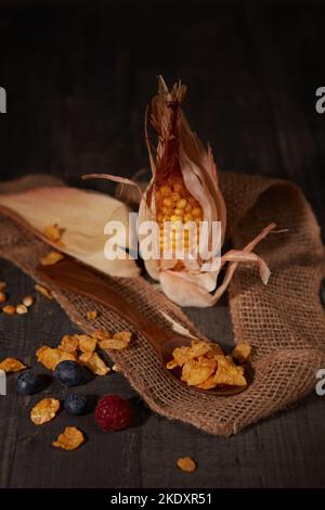 Frischer Mais und Cornflakes in der Nähe von reifen Waldbeeren und Streifen aus Sackleinen Stoff auf Holztisch im dunklen Raum platziert Stockfoto