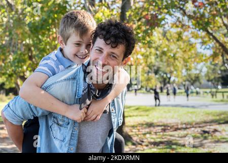 Positiver Vater lächelt und schaut in die Kamera, während er seinem Sohn am Herbstwochenende im Park auf einem gepflasterten Weg eine Huckepack-Fahrt gibt Stockfoto