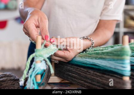 Crop unkenntlich Frau in Freizeitkleidung mit Maniküre Webteppich aus bunten Fäden auf Webstuhl in der Werkstatt Stockfoto