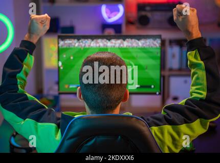 Junger Mann, der zu Hause Sport im fernsehen sieht und ein schreiendes, fröhliches Fußballspiel feiert. Stockfoto