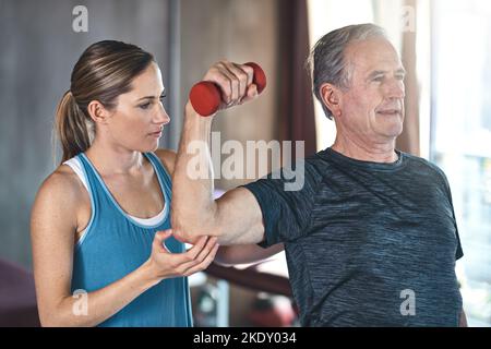 Bewegung ist der Schlüssel zu einem gesunden Geist, Herz und Körper. Ein älterer Mann mit Gewichten mit Hilfe eines Physiotherapeuten. Stockfoto