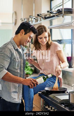 Eine attraktive junge Frau, die Wein trinkt, während sie ihrem Mann beim Kochen zuschaut. Stockfoto