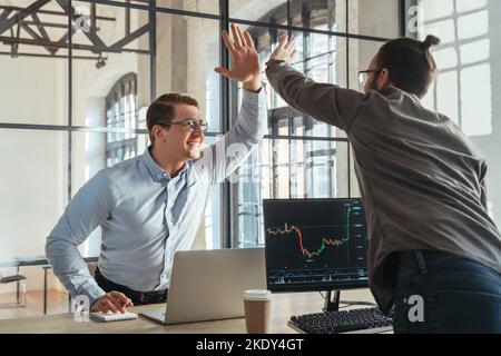 Mitarbeiter-Trader, die sich gegenseitig hohe fünf geben, feiern einen erfolgreichen Deal am Aktienmarkt vor Computern mit Candlestick-Charts. IPO-Gewinn. Kryptowährungsinvestoren Stockfoto