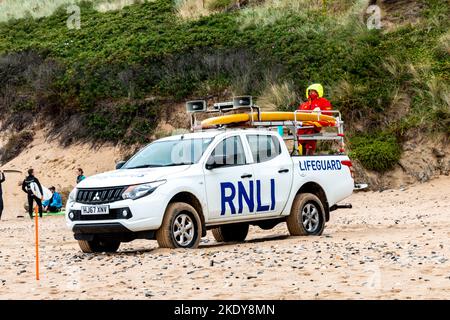 An einem kalten Tag trägt der RNLI-Rettungsschwimmer einen großen Mantel mit der Haube nach oben, während er die Uhr von der Rückseite seines Pick-up-Trucks, Fistral Beach, Newquay, hält. Stockfoto