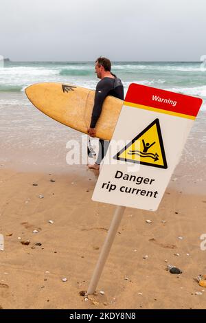 Am Fistral Beach, Newquay, läuft ein Surfer an einem Schild vorbei, das warnt, dass es eine gefährliche Stromreißströmung gibt. Stockfoto