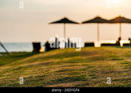 Verschwommenes Bild kleine Grasberge mit dem Hintergrund von Menschen, die auf Strandliegen unter Sonnenschirmen sitzen, Sonnenuntergangszeit mit goldenem Himmel und Meer. Stockfoto