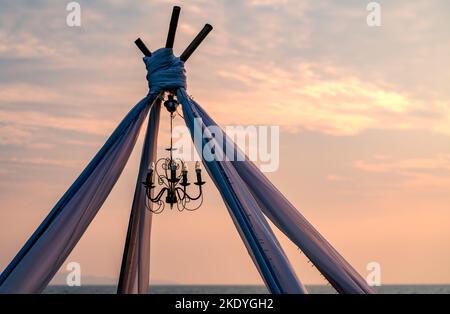 Nahaufnahme eines alten Kronleuchters mit Lampen, die unter der Hochzeitshütte am Strand bei Sonnenuntergang hängen Stockfoto