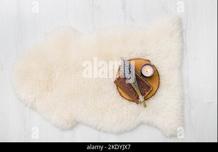 Flach Lay-Ansicht von weißen warmen und gemütlichen ganzen echten dekorativen Schaffell Teppich auf weißem Holzbrett Boden, oben ist Buch, Blumen und Kerze. Speicherplatz kopieren. Stockfoto