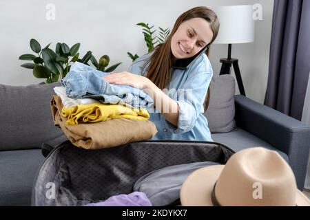 Glückliche Millennial Dame diskutieren Koffer Verpackung Kleidung Organisation auf Smartphone sitzen auf der Couch. Positive Mädchen reden Handy-Liste machen Stockfoto