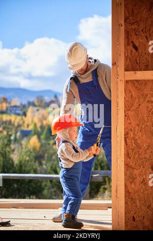 Vater mit Kleinkind Sohn Gebäude Holzrahmen Haus. Junge hilft seinem Vater, schlägt Nagel auf die Planke auf der Baustelle, trägt Helm und blaue Overalls an sonnigen Tagen. Zimmerei- und Familienkonzept. Stockfoto